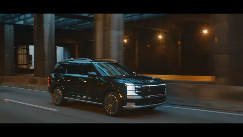 John Krasinski driving the Hyundai Palisade with a city skyline in the background